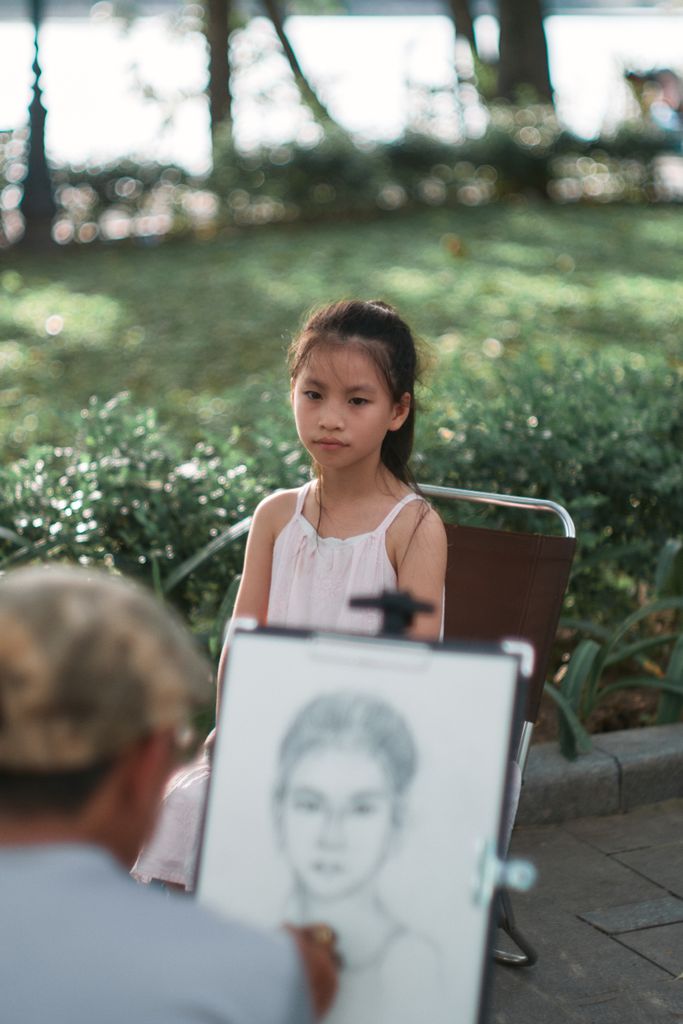 A young girl posing for a sketch artist in a park, captured in natural lighting.