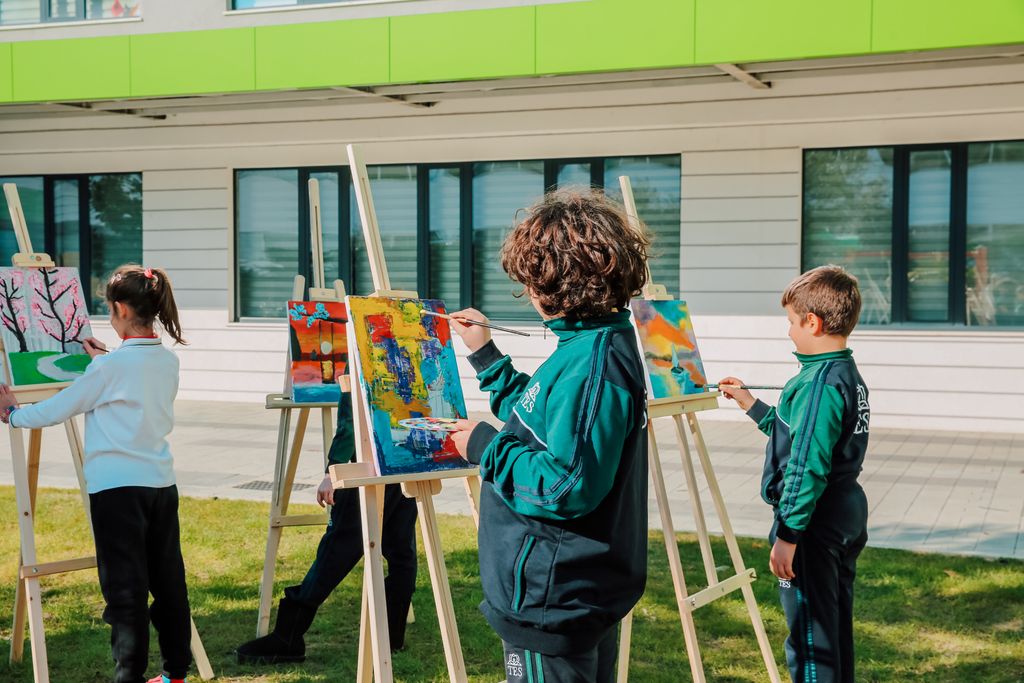 Children painting colorful designs on easels outdoors in school courtyard.