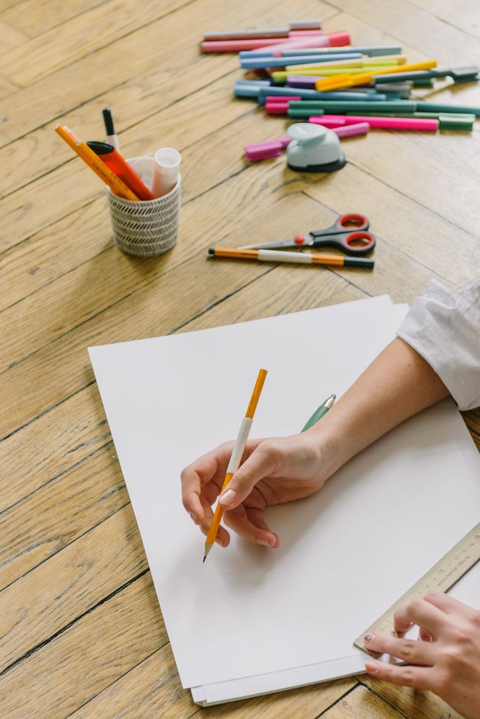 Close-up of hands drawing on white paper surrounded by art supplies, including pencils, markers, and scissors, on a wooden floor.