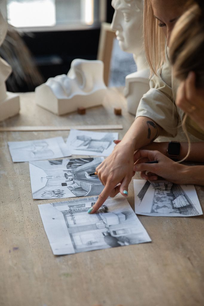 Two artists reviewing pencil sketches on a wooden table in a studio setting with sculptures.